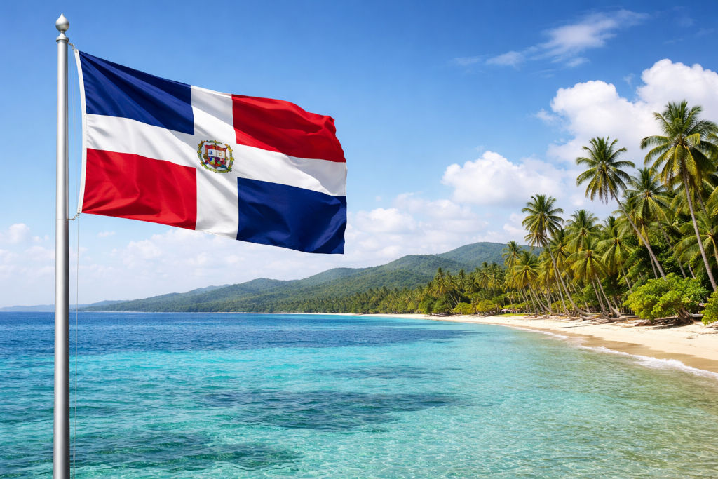 Nationalflagge der Dominikanischen Republik mit rotem und blauem Kreuz sowie Staatswappen in der Mitte, links am Fahnenmast wehend, vor karibischer Küstenlandschaft mit türkisfarbenem Meer, hellem Sandstrand, Palmen, grünen Hügeln und blauem Himmel mit Wolken im 16:9‑Format