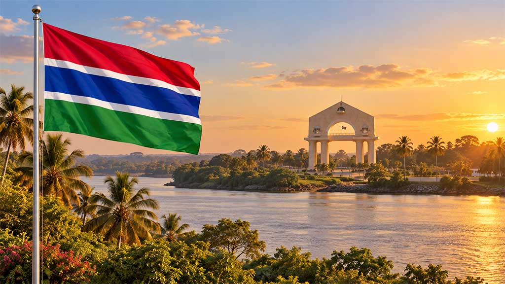 Flagge von Gambia am silbernen Fahnenmast vor dem Gambia River, westafrikanischer Savannenlandschaft, Palmen und dem bekannten Arch 22 in Banjul bei warmem Golden-Hour-Licht.