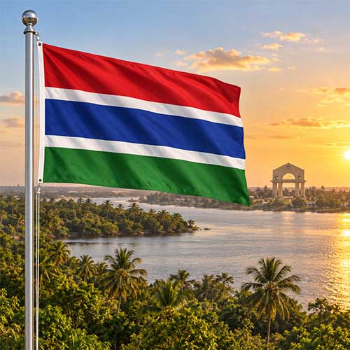 Gambias Nationalflagge am silbernen Fahnenmast vor dem Gambia River mit Palmen, Savannenlandschaft und dem Arch 22 in Banjul im warmen Abendlicht.