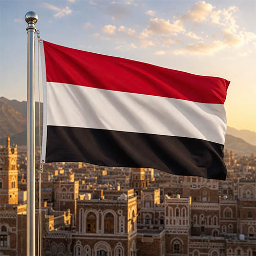 Flagge des Jemen vor der historischen Altstadt von Sanaa mit ihren Lehmziegelhäusern und dekorativen weißen Fassaden in warmer Abendstimmung.