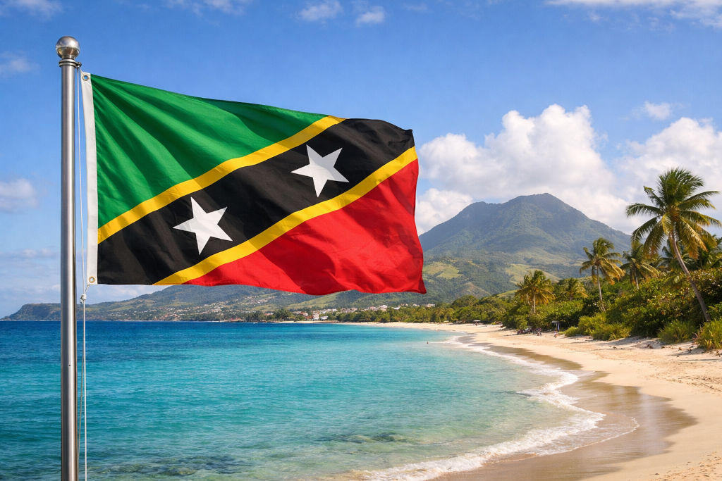 Nationalflagge von St. Kitts und Nevis an einem Fahnenmast wehend; im Hintergrund tropische Küstenlandschaft mit türkisfarbenem Meer, hellem Sandstrand, Palmen und bewaldetem Vulkanberg unter blauem Himmel, im Format 16:9